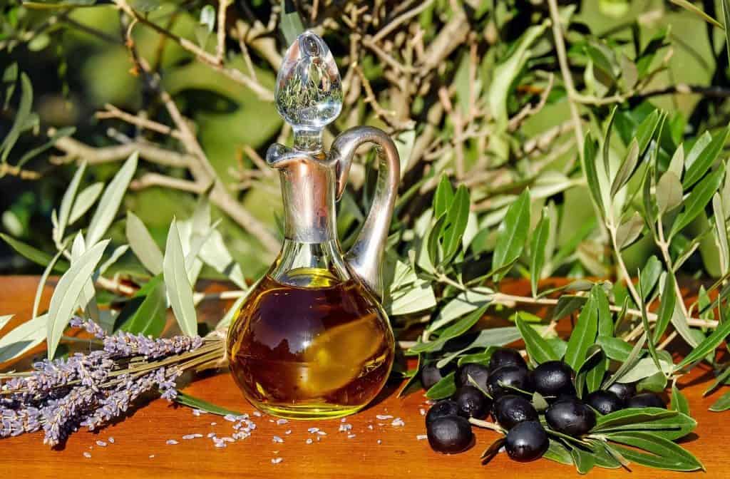 Olivenöl in einer Karaffe draußen auf einem Tisch mit Oliven und Ästen.